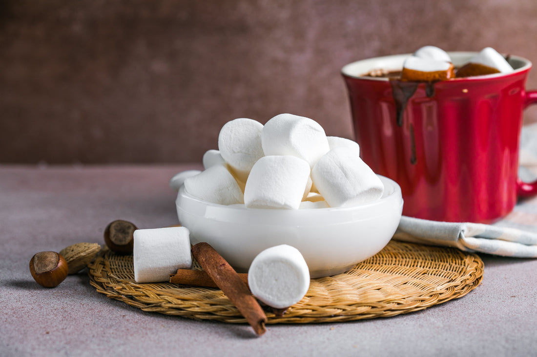 A bowl of fluffy gourmet marshmallows ready to be added to a steaming cup of hot chocolate. Cinnamon sticks and hazelnuts complete the cozy winter scene.